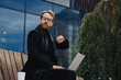 © YURII MASLAK - Successful businessman on the background of the business center. Red-haired young guy, in a stylish coat, wireless headphones with a laptop.