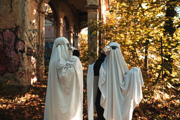 A funny image of two people in ghost costumes and sunglasses holding a cup with a drink in an abandoned building