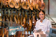 © JackF - Cheerful caucasian woman in uniform working in jamoneria, standing at showcase and smiling.