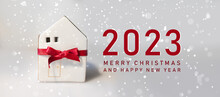 Merry Christmas And 2023 Happy New Year Horizontal Banner With Small Toy Model House Wrapped In Red Satin Ribbon On A White Background. Miniature White Toy House Model On Color Background.
