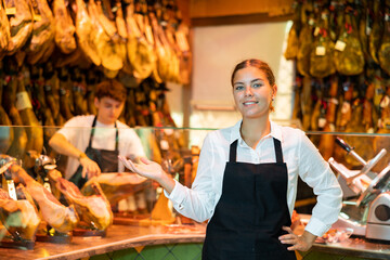 Successful smiling young woman, butcher shop owner, standing in apron near glass counter, greeting visitors and offering delicious Iberian jamon