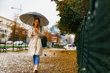 Wall Mural - Happy caucasian female with an umbrella on rainy day, walks down a street looking at her phone