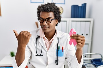 Canvas Print - African doctor man holding pink cancer ribbon pointing thumb up to the side smiling happy with open mouth