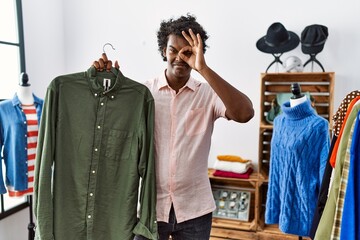 Canvas Print - African man with curly hair holding shirt from clothing rack at retail shop smiling happy doing ok sign with hand on eye looking through fingers