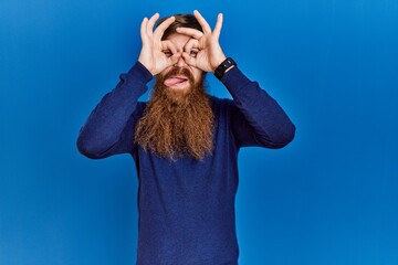 Canvas Print - Redhead man with long beard wearing casual blue sweater over blue background doing ok gesture like binoculars sticking tongue out, eyes looking through fingers. crazy expression.