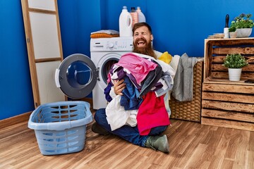 Canvas Print - Redhead man with long beard putting dirty laundry into washing machine winking looking at the camera with sexy expression, cheerful and happy face.