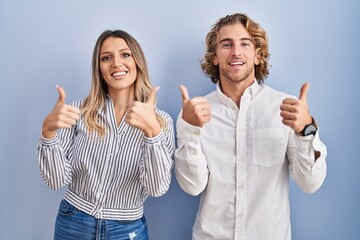 Wall Mural - Young couple standing over blue background success sign doing positive gesture with hand, thumbs up smiling and happy. cheerful expression and winner gesture.