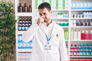 Poster - Handsome hispanic man working at pharmacy drugstore smelling something stinky and disgusting, intolerable smell, holding breath with fingers on nose. bad smell