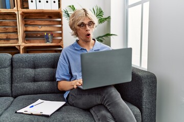 Canvas Print - Young man working using computer laptop sitting on the sofa scared and amazed with open mouth for surprise, disbelief face