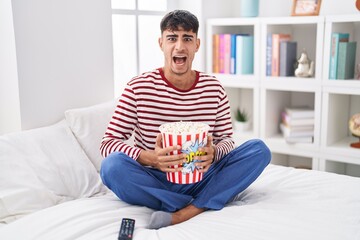 Wall Mural - Young hispanic man eating popcorn sitting on the bed watching a movie angry and mad screaming frustrated and furious, shouting with anger. rage and aggressive concept.