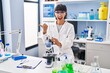 © Krakenimages.com - Young hispanic woman working at scientist laboratory pointing thumb up to the side smiling happy with open mouth