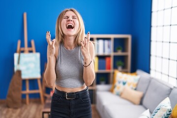 Sticker - Blonde caucasian woman standing at living room crazy and mad shouting and yelling with aggressive expression and arms raised. frustration concept.