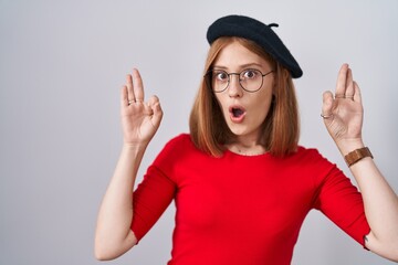 Canvas Print - Young redhead woman standing wearing glasses and beret looking surprised and shocked doing ok approval symbol with fingers. crazy expression