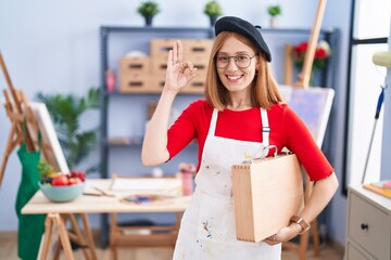 Poster - Young redhead woman at art studio holding art case smiling positive doing ok sign with hand and fingers. successful expression.