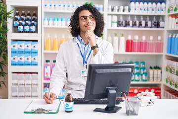 Sticker - Hispanic man with curly hair working at pharmacy drugstore with hand on chin thinking about question, pensive expression. smiling and thoughtful face. doubt concept.