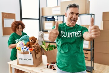 Poster - Middle age man wearing volunteer t shirt at donations stand approving doing positive gesture with hand, thumbs up smiling and happy for success. winner gesture.