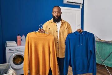 Wall Mural - African american man holding clean clothes on hangers at laundry room making fish face with mouth and squinting eyes, crazy and comical.