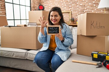 Canvas Print - Young hispanic woman holding blackboard with new home text doing ok sign with fingers, smiling friendly gesturing excellent symbol