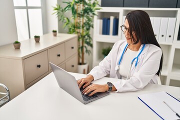 Wall Mural - Young hispanic woman wearing doctor uniform using laptop working at clinic