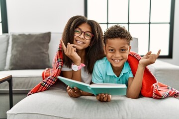 Wall Mural - Two siblings lying on the sofa reading a book with a big smile on face, pointing with hand finger to the side looking at the camera.