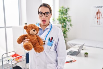 Poster - Young hispanic pediatrician woman holding teddy bear at the clinic scared and amazed with open mouth for surprise, disbelief face