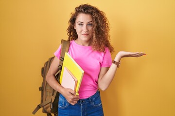 Sticker - Young caucasian woman wearing student backpack and holding books smiling cheerful presenting and pointing with palm of hand looking at the camera.