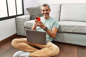 Canvas Print - Middle age grey-haired man using smartphone and laptop sitting on floor at home