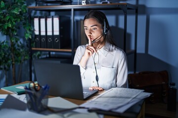 Poster - Young brunette woman wearing call center agent headset working late at night asking to be quiet with finger on lips. silence and secret concept.