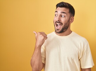 Wall Mural - Handsome hispanic man standing over yellow background smiling with happy face looking and pointing to the side with thumb up.