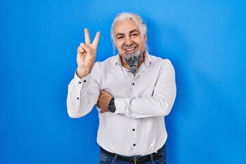 Wall Mural - Middle age man with grey hair standing over blue background smiling with happy face winking at the camera doing victory sign with fingers. number two.