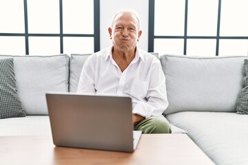 Sticker - Senior man using laptop at home sitting on the sofa puffing cheeks with funny face. mouth inflated with air, crazy expression.