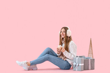 Sticker - Young woman with glass cup of cocoa and Christmas presents sitting on pink background