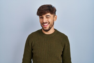 Wall Mural - Arab man with beard standing over blue background winking looking at the camera with sexy expression, cheerful and happy face.