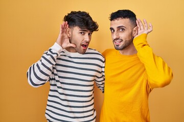 Canvas Print - Young hispanic gay couple standing over yellow background smiling with hand over ear listening an hearing to rumor or gossip. deafness concept.