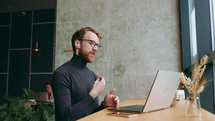 A young red-haired guy, a programmer or businessman in glasses, is sitting in a stylish cafe at a laptop and is talking through a webcam. Freelancer works remotely. Online communication.