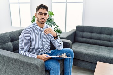 Canvas Print - Young psychologist man at consultation office relaxed with serious expression on face. simple and natural looking at the camera.