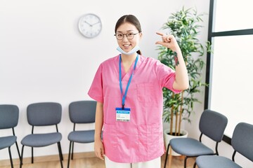 Canvas Print - Young asian nurse woman at medical waiting room smiling and confident gesturing with hand doing small size sign with fingers looking and the camera. measure concept.