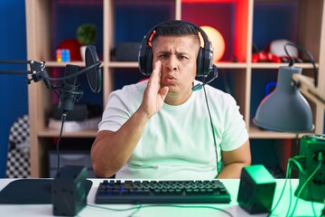 Poster - Young hispanic man playing video games hand on mouth telling secret rumor, whispering malicious talk conversation