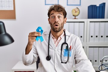 Canvas Print - Young hispanic doctor man holding blue ribbon scared and amazed with open mouth for surprise, disbelief face