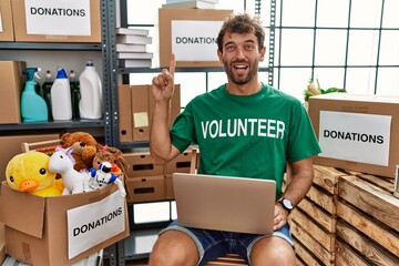 Canvas Print - Young handsome man wearing volunteer t shirt using laptop pointing finger up with successful idea. exited and happy. number one.