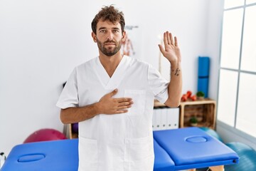 Poster - Young handsome physiotherapist man working at pain recovery clinic swearing with hand on chest and open palm, making a loyalty promise oath