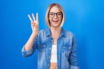 Sticker - Young caucasian woman standing over blue background showing and pointing up with fingers number three while smiling confident and happy.