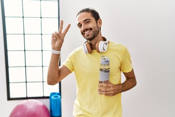 Canvas Print - Young hispanic man wearing sportswear and drinking water at the gym smiling looking to the camera showing fingers doing victory sign. number two.