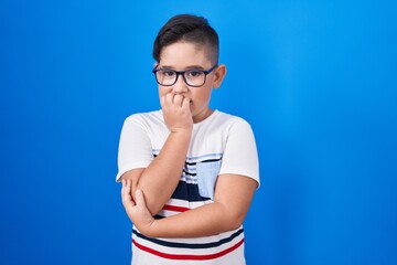 Canvas Print - Young hispanic kid standing over blue background looking stressed and nervous with hands on mouth biting nails. anxiety problem.