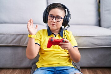 Sticker - Young hispanic kid playing video game holding controller wearing headphones waiving saying hello happy and smiling, friendly welcome gesture