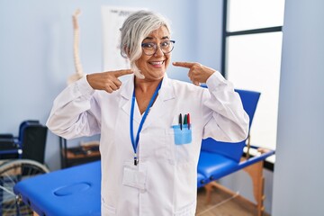 Wall Mural - Middle age woman with grey hair working at pain recovery clinic smiling cheerful showing and pointing with fingers teeth and mouth. dental health concept.