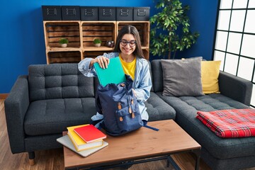 Poster - Young hispanic woman student holding books of backpack sitting on sofa at home