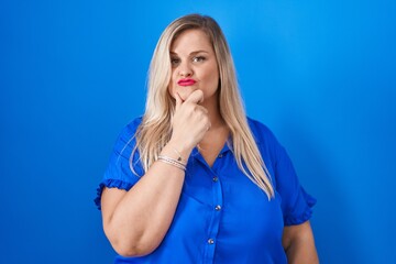 Canvas Print - Caucasian plus size woman standing over blue background looking confident at the camera with smile with crossed arms and hand raised on chin. thinking positive.