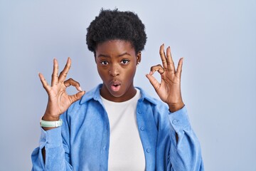 Sticker - African american woman standing over blue background looking surprised and shocked doing ok approval symbol with fingers. crazy expression