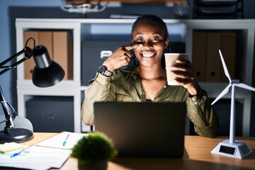 Poster - African woman working using computer laptop at night pointing with hand finger to face and nose, smiling cheerful. beauty concept